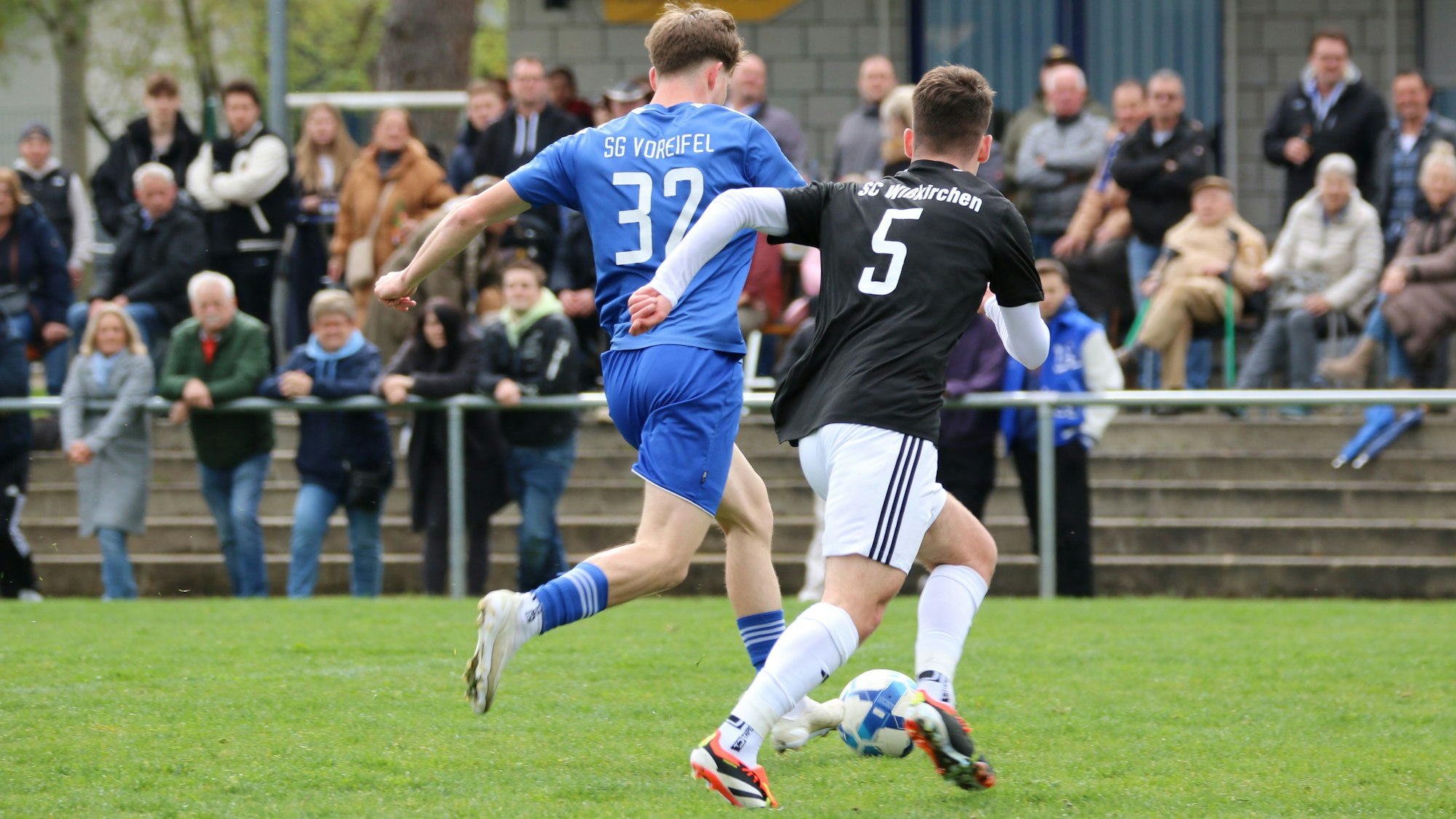 Zwei Spieler der SG Voreifel und des SC Wißkirchen im Laufduell um den Ball. Im Hintergrund sind Zuschauer des Fußballspiels auf einer Stehstufenanlage zu sehen.