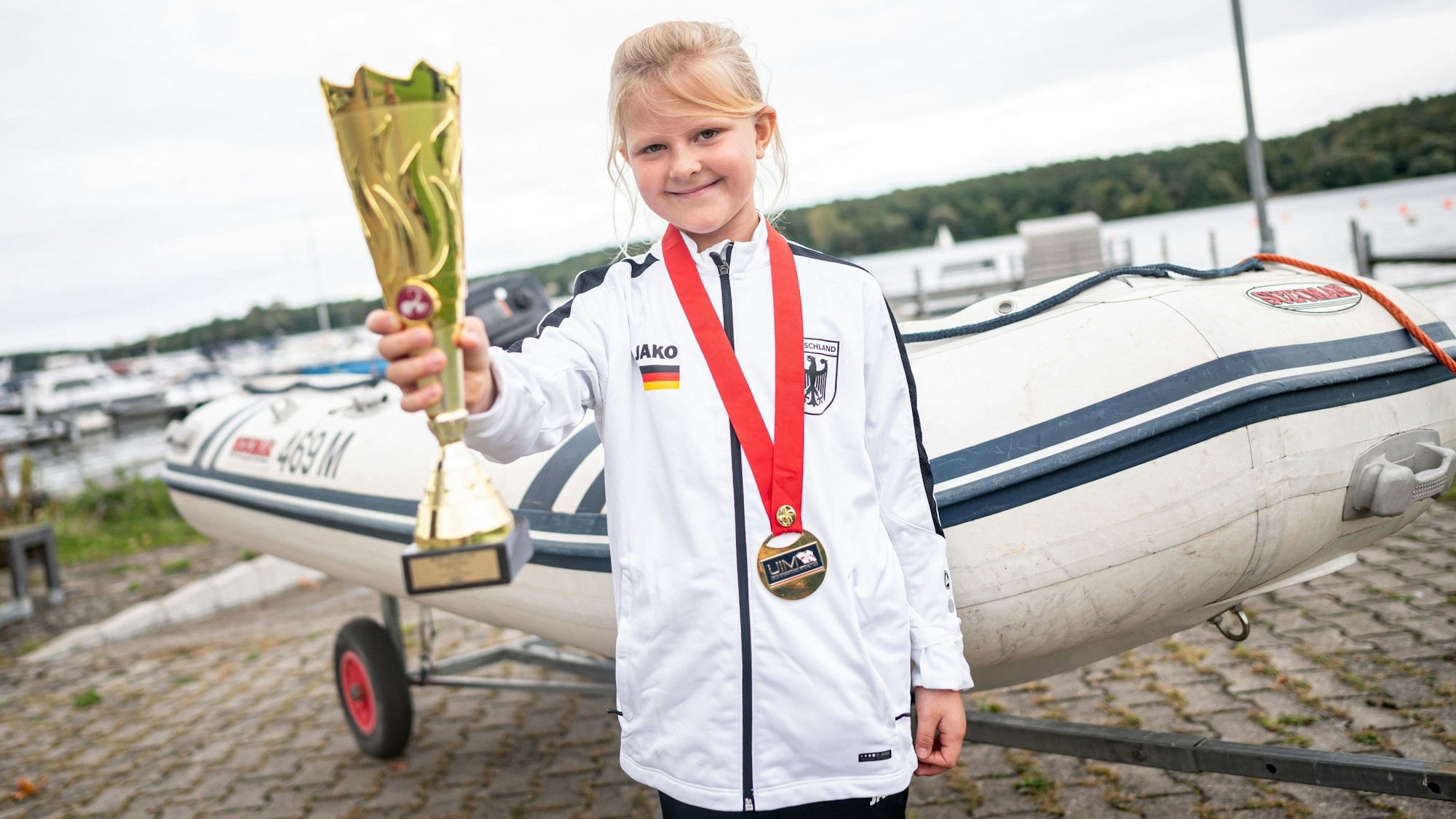 PRODUKTION - 25.09.2024, Brandenburg, Beetzsee/Brielow: Jana Pietack, siebenjährige Weltmeisterin im Schlauchbootfahren, steht mit Pokal und Medaille vor ihrem Schlauchboot. (zu dpa: «Siebenjährige Berlinerin ist Schlauchboot-Weltmeisterin») Foto: Fabian Sommer/dpa +++ dpa-Bildfunk +++