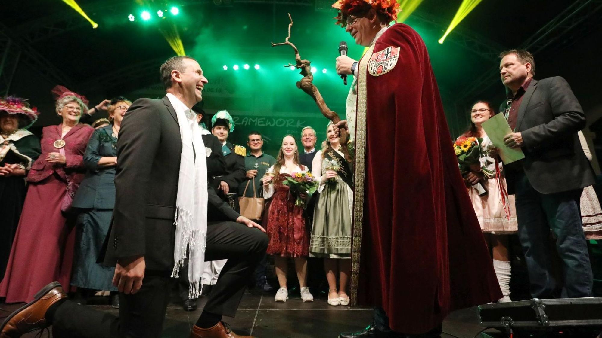 Ein Mann im schwarzen Anzug kniet, vor ihm der Darsteller des Weingottes Bacchus in roter Robe mit Weinstock und Mikrofon.