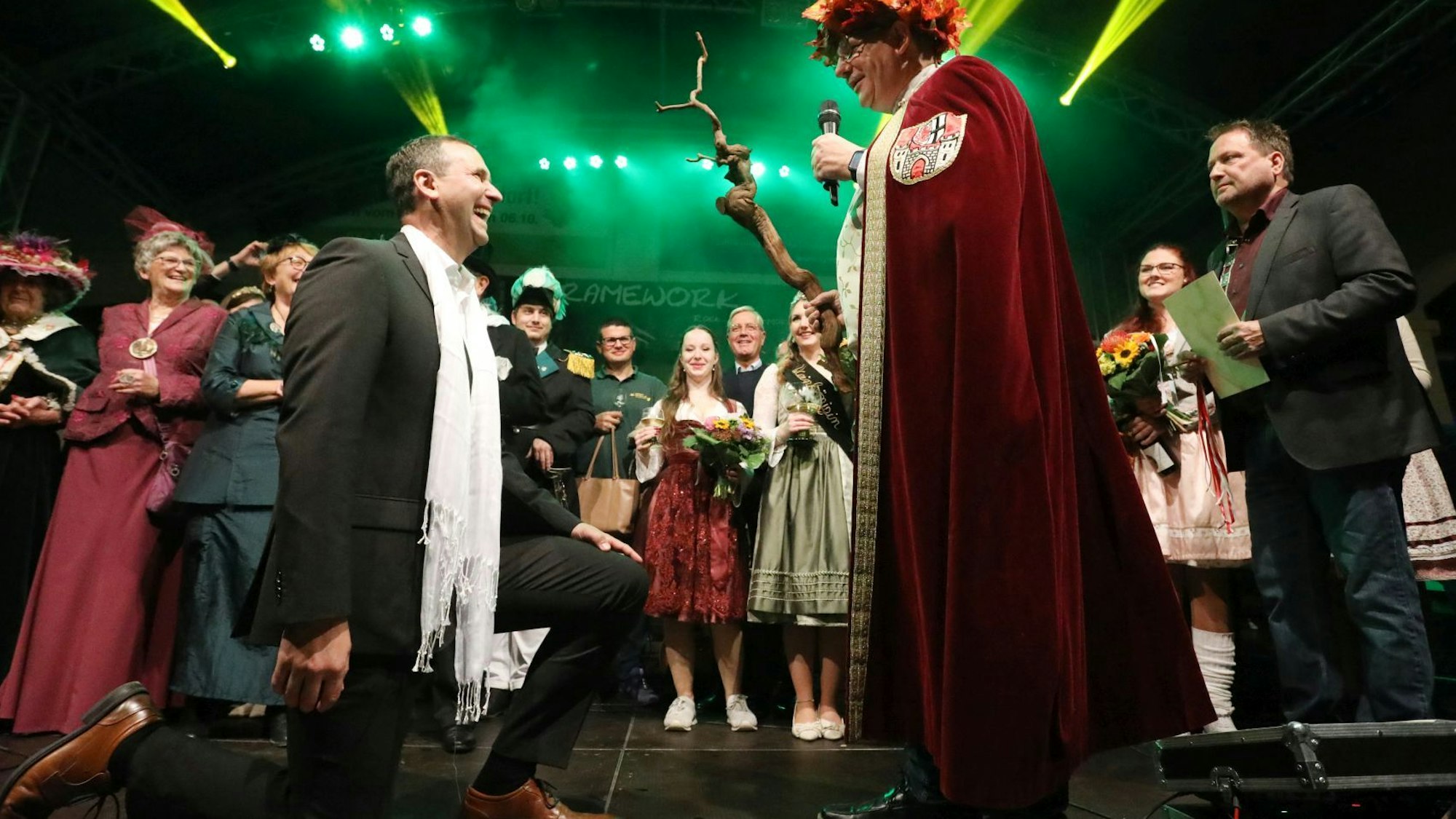 Ein Mann im schwarzen Anzug kniet, vor ihm der Darsteller des Weingottes Bacchus in roter Robe mit Weinstock und Mikrofon.