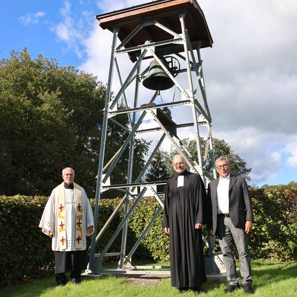 Drei Männer, darunter zwei Geistliche, stehen vor einem neuen Glockenturm.