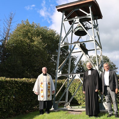Drei Männer, darunter zwei Geistliche, stehen vor einem neuen Glockenturm.