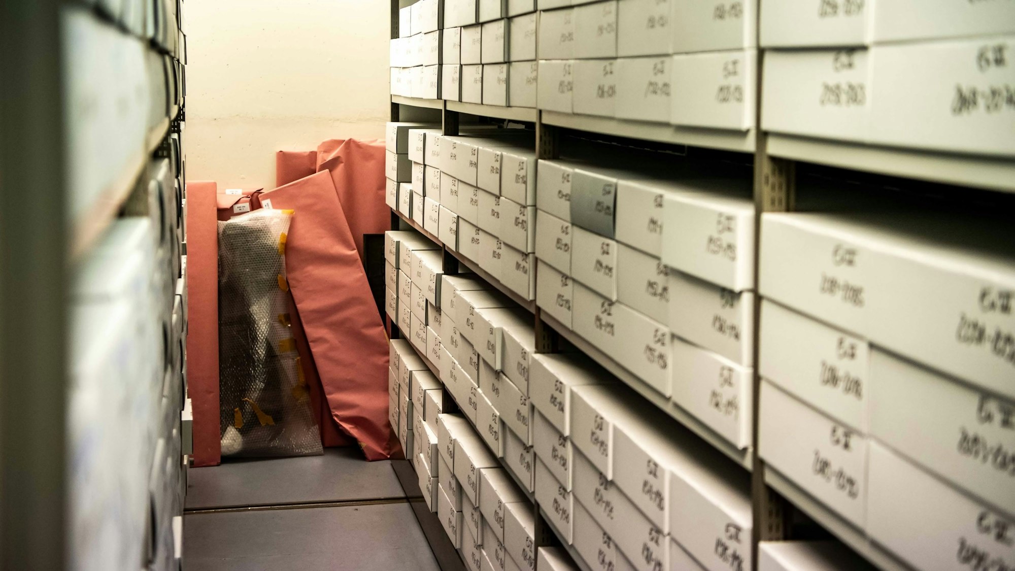 Ein enger Gang, in den Regalen zu beiden Seiten stapeln sich zahlreiche beschriftete Schachteln, in denen Archivalien befinden.
