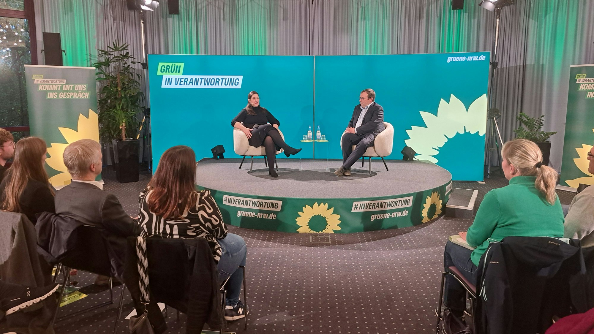 Grünen NRW-Vorsitzende Yazgülü Zeybek und Minister Oliver Krischer im Gespräch, sie sitzen auf einem halbrunden Podium.