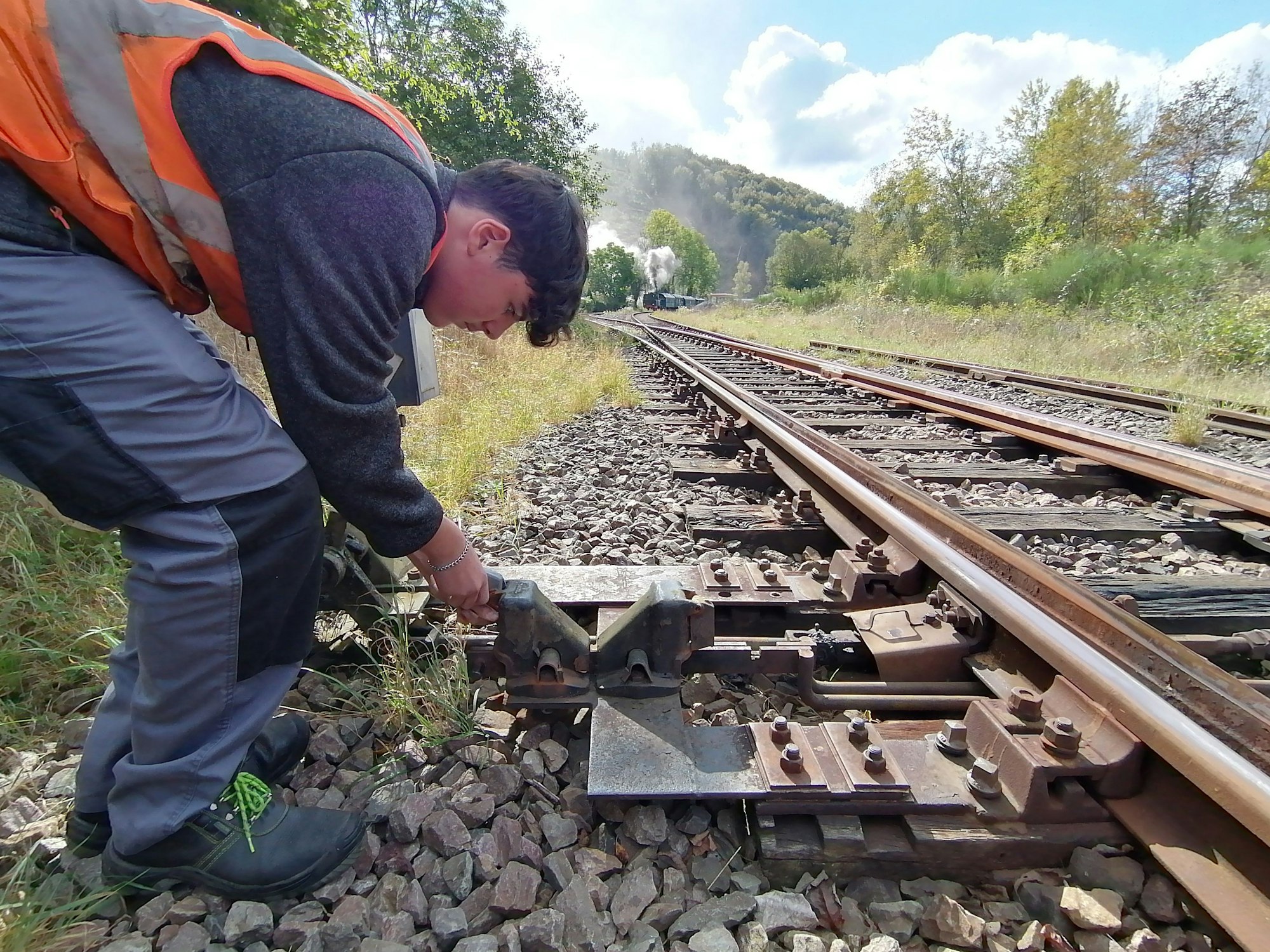Ein junger Mann mit Warnweste steht über dem Hebel einer Weiche an einem Bahngleis gebeugt.