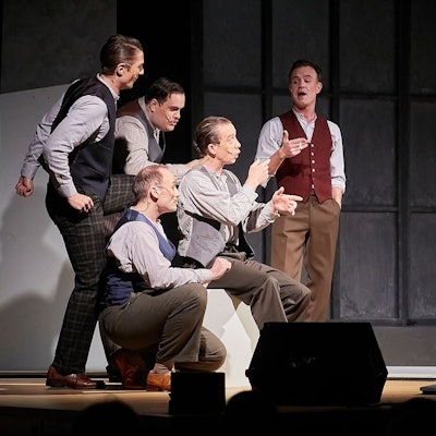 Eine Gruppe von fünf Männern, die im Stil der 1930er Jahre gekleidet sind, stehen gestikulierend auf der Bühne.