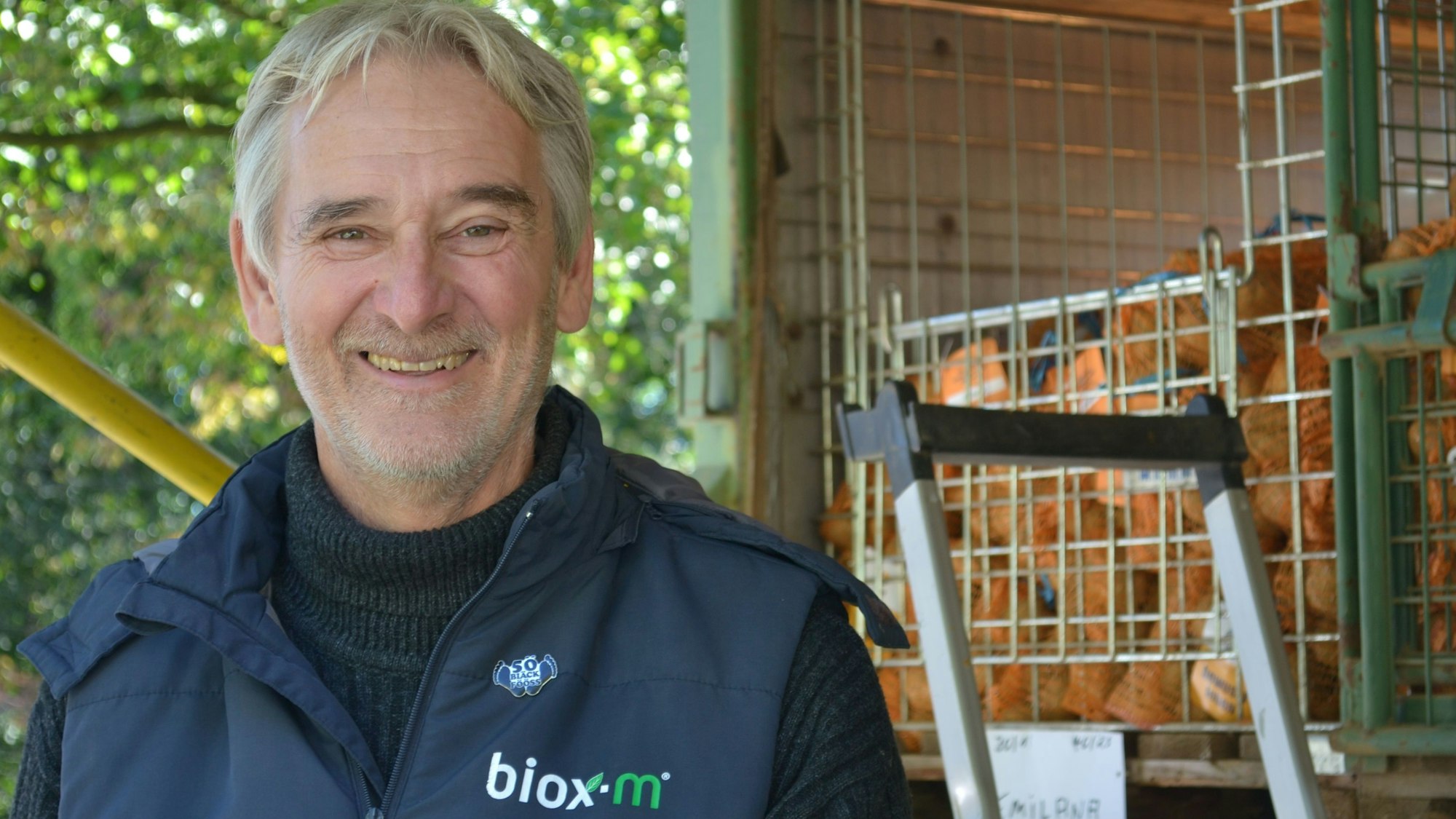 Ein grauhaariger Mann lacht in die Kamera, hinter ihm eine Gitterbox mit Kartoffeltüten.