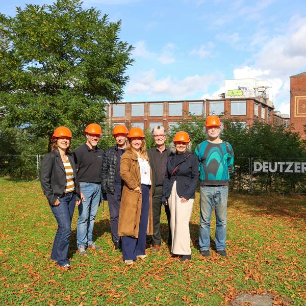 Einige Frauen und Männer mit roten Helmen auf dem Kopf stehen vor einem alten Industriegebäude.