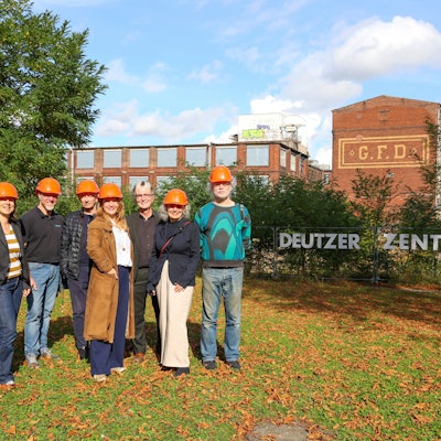 Einige Frauen und Männer mit roten Helmen auf dem Kopf stehen vor einem alten Industriegebäude.