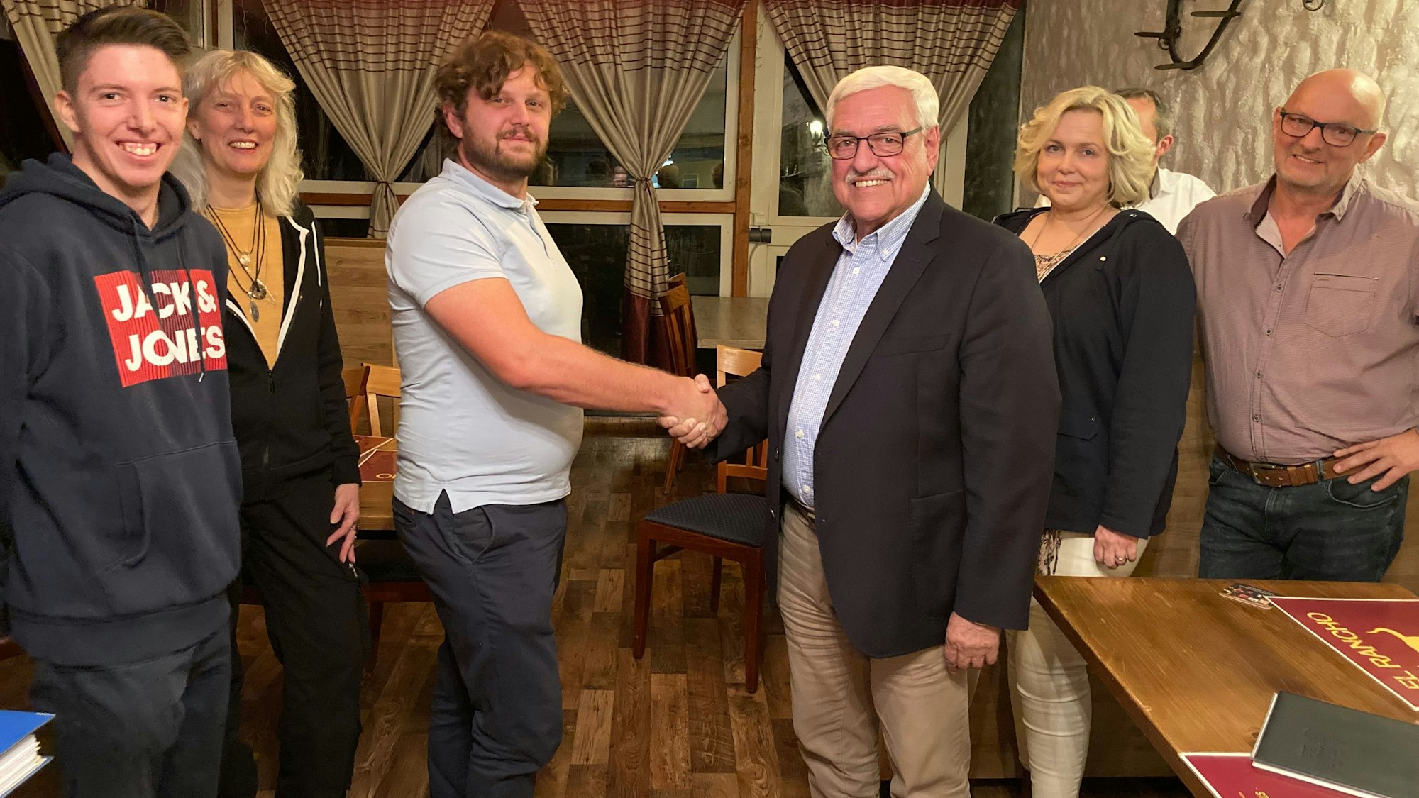 Ein Mann schüttelt einem älteren Herrn die Hand, während beide in die Kamera schauen.