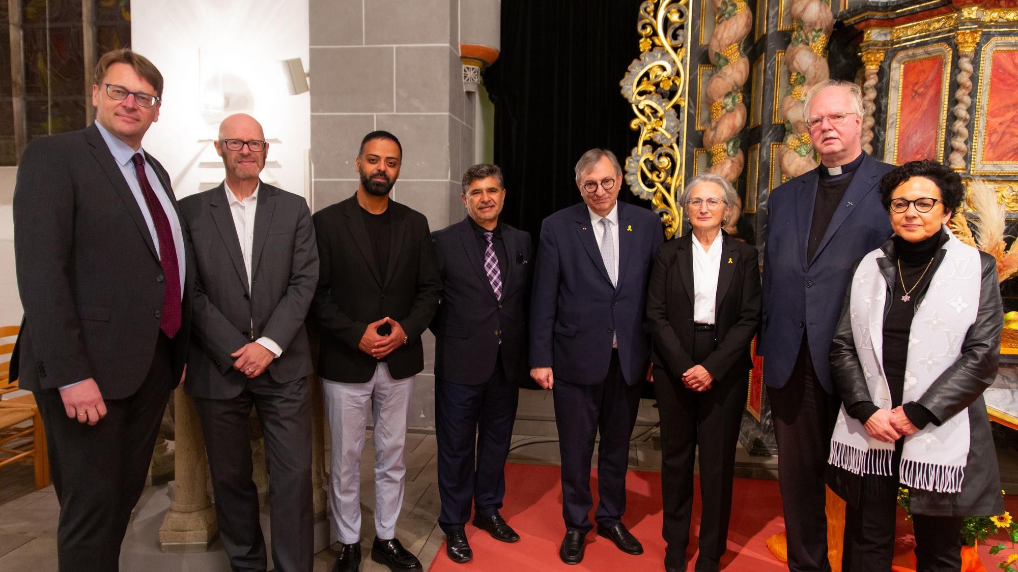 Zahlreiche Männer und einer Frau unterschiedlicher Religionen stehen bei einer Gedenkstunde in einer Kirche beisammen.