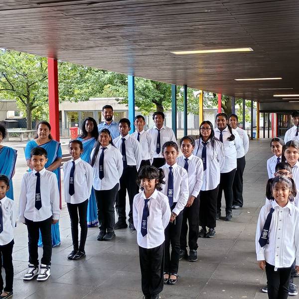 Auf dem Bild sind die Schülerinnen und Schüler zu sehen, die sich in drei Reihen aufgestellt haben, bevor es in die Klassen geht. Sie tragen Schuluniformen. Links und rechts neben ihnen haben sich die Lehrerinnen und Lehrer aufgestellt. Die Lehrerinnen tragen blaue Sahrees.
