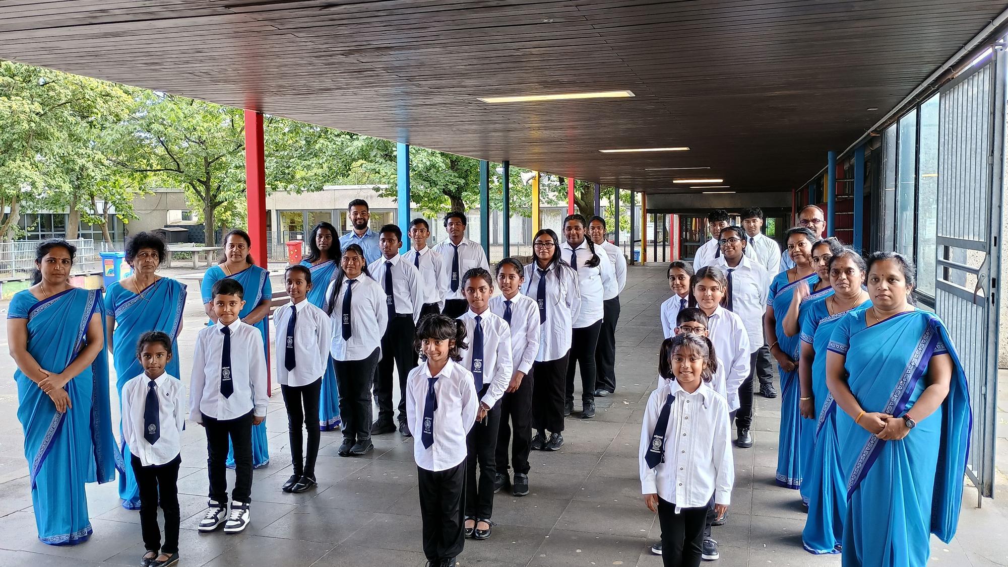 Auf dem Bild sind die Schülerinnen und Schüler zu sehen, die sich in drei Reihen aufgestellt haben, bevor es in die Klassen geht. Sie tragen Schuluniformen. Links und rechts neben ihnen haben sich die Lehrerinnen und Lehrer aufgestellt. Die Lehrerinnen tragen blaue Sahrees.