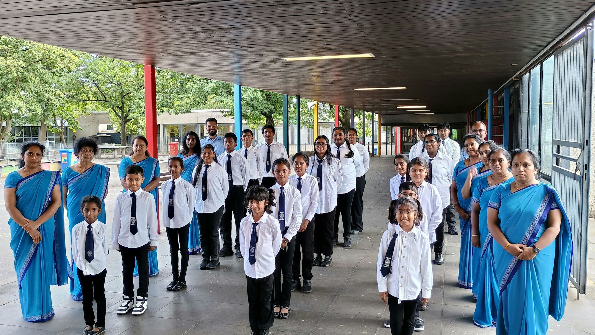 Auf dem Bild sind die Schülerinnen und Schüler zu sehen, die sich in drei Reihen aufgestellt haben, bevor es in die Klassen geht. Sie tragen Schuluniformen. Links und rechts neben ihnen haben sich die Lehrerinnen und Lehrer aufgestellt. Die Lehrerinnen tragen blaue Sahrees.