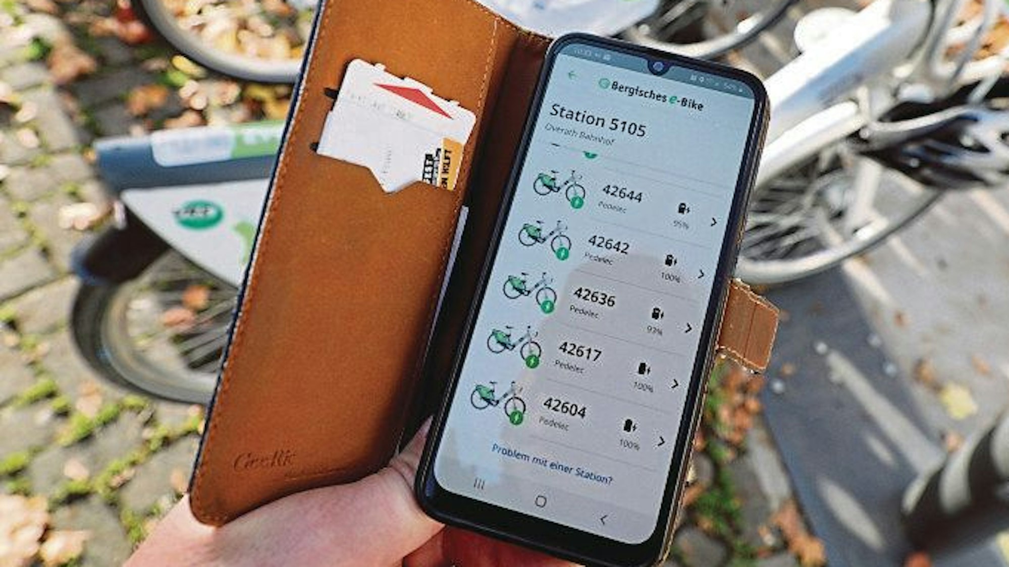 Ein Handy mit der App des „Bergischen E-Bike“ ist vor abgestellten E-Bikes des Verleihsystems zu sehen.