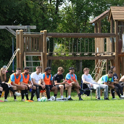 Das Bild zeigt eine Reservebank mit Fußballspielern. Im Hintergrund ist ein Spielplatz zu erkennen.