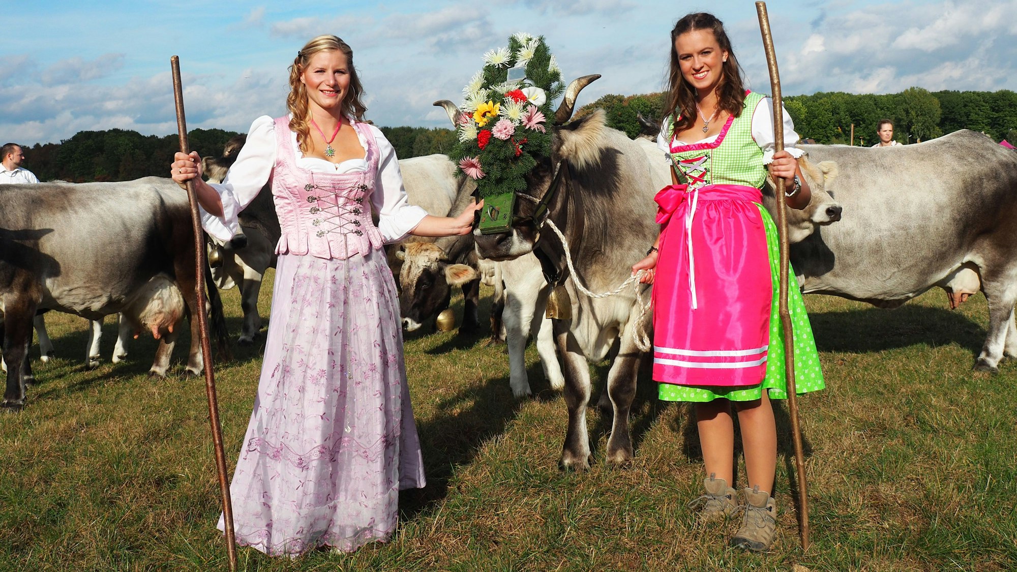 Zwei Frauen im Dirndl stehen vor einer Herde Kühe, eine ist prachtvoll geschmückt und hat eine Glocke um den Hals