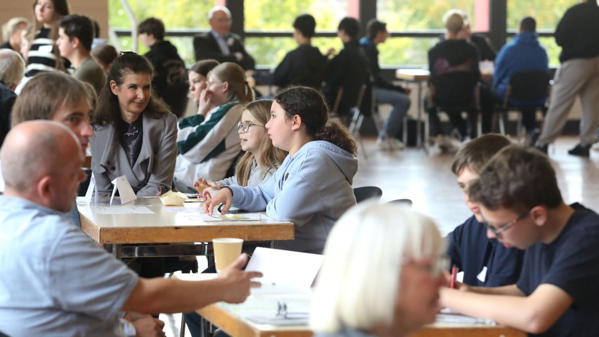 Schüler und Erwachsene sitzen an mehreren Tischen, teilweise machen sie sich Notizen.