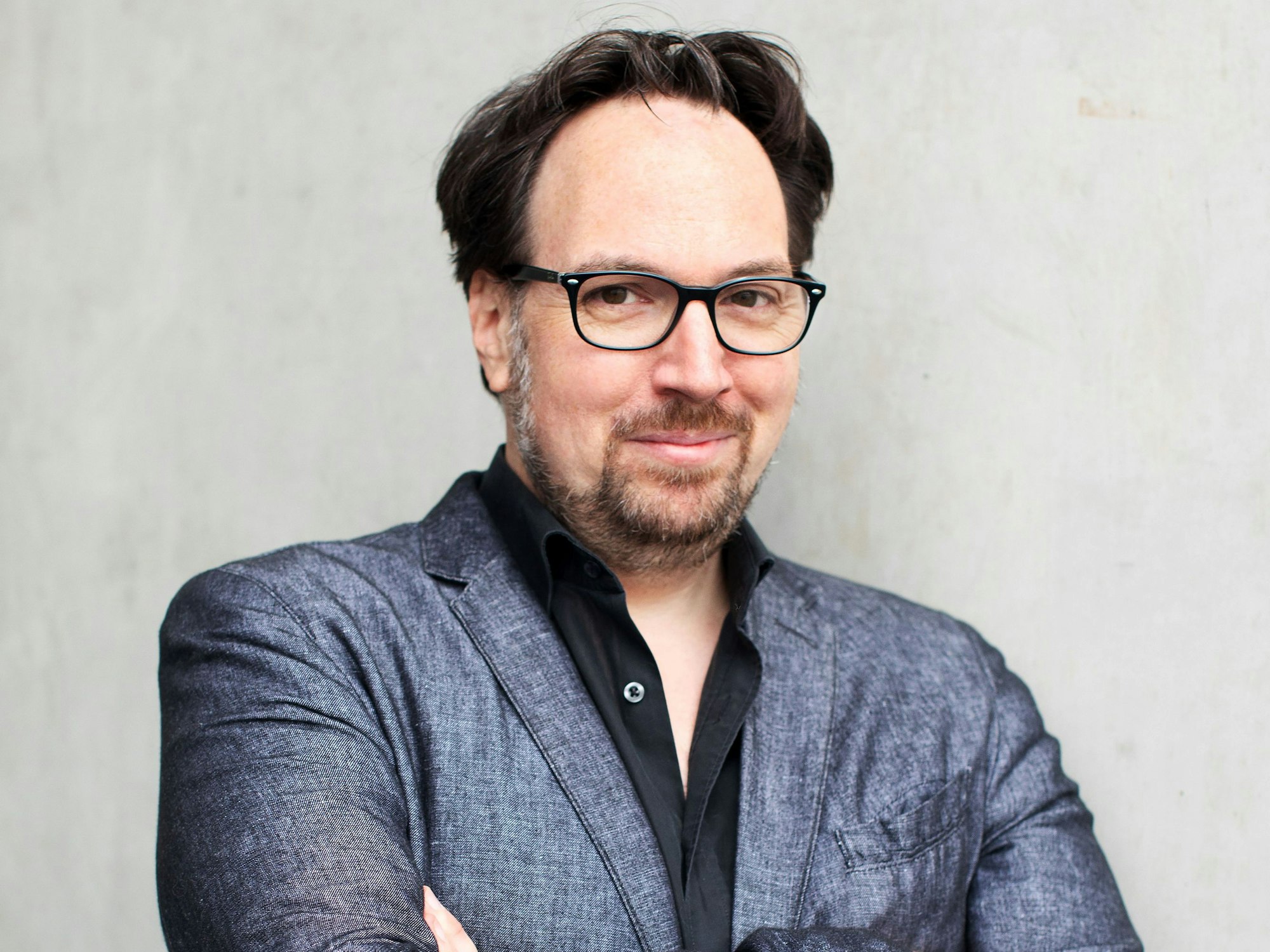 Der Autor trägt eine Brille, einen Bart und ein dunkelgraues Sakko. Darunter ein schwarzes Hemd.
