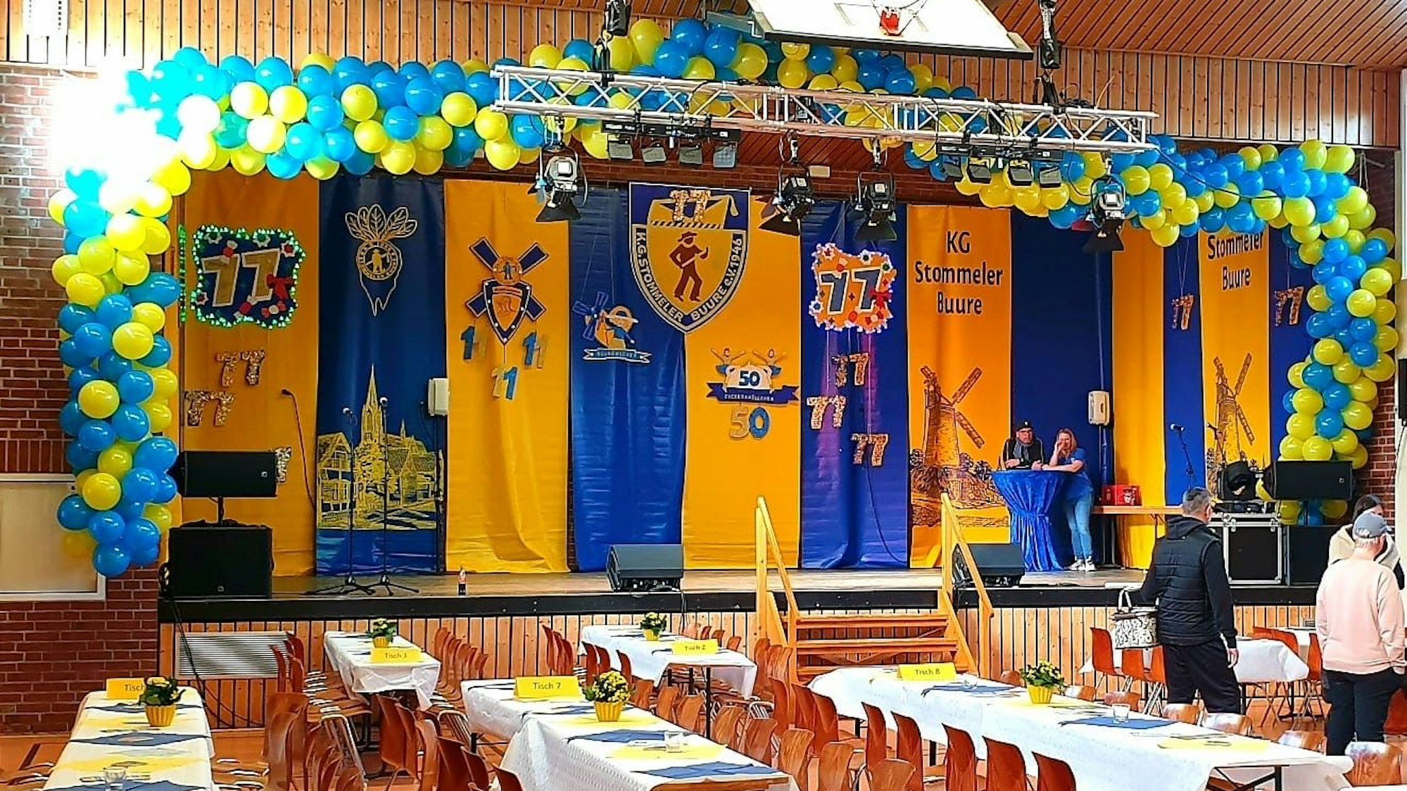 Blick in eine Veranstaltungshalle mit einer Bühne, davor stehen Tische.