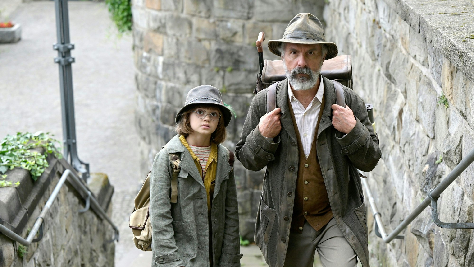 Auf dem Bild sind Christoph Maria Herbst und Yuna Bennett in ihren Rollen als gemeinsame Buchspazierer zu sehen. Sie gehen eine steinerne Treppe hoch. Die beiden tragen Hüte und Jacken.