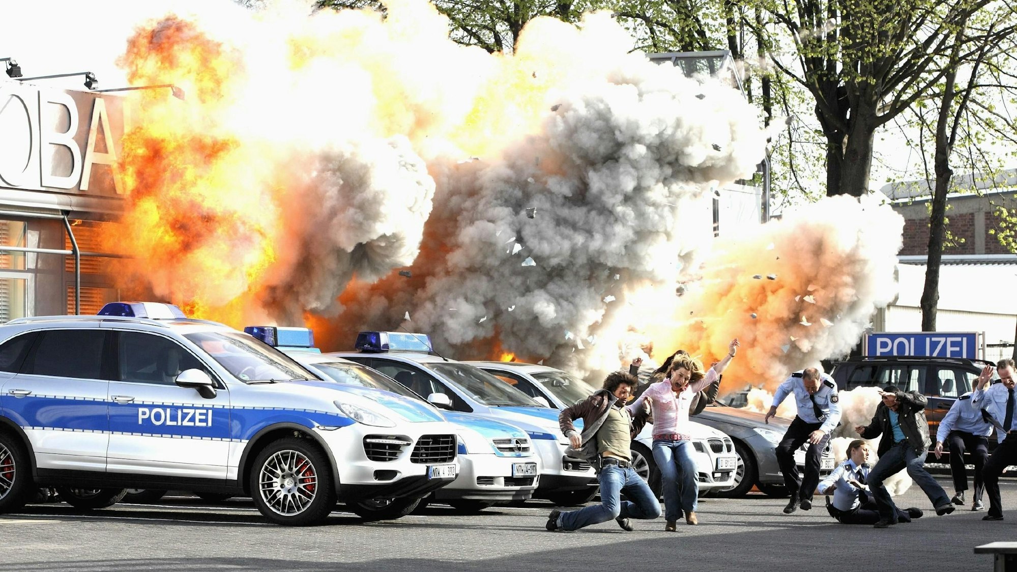 Das Foto zeigt eine Filmexplosion in der Kulisse der Autobahnpolizeiwache. Davor mehrere Polizeiautos und flüchtende Menschen.