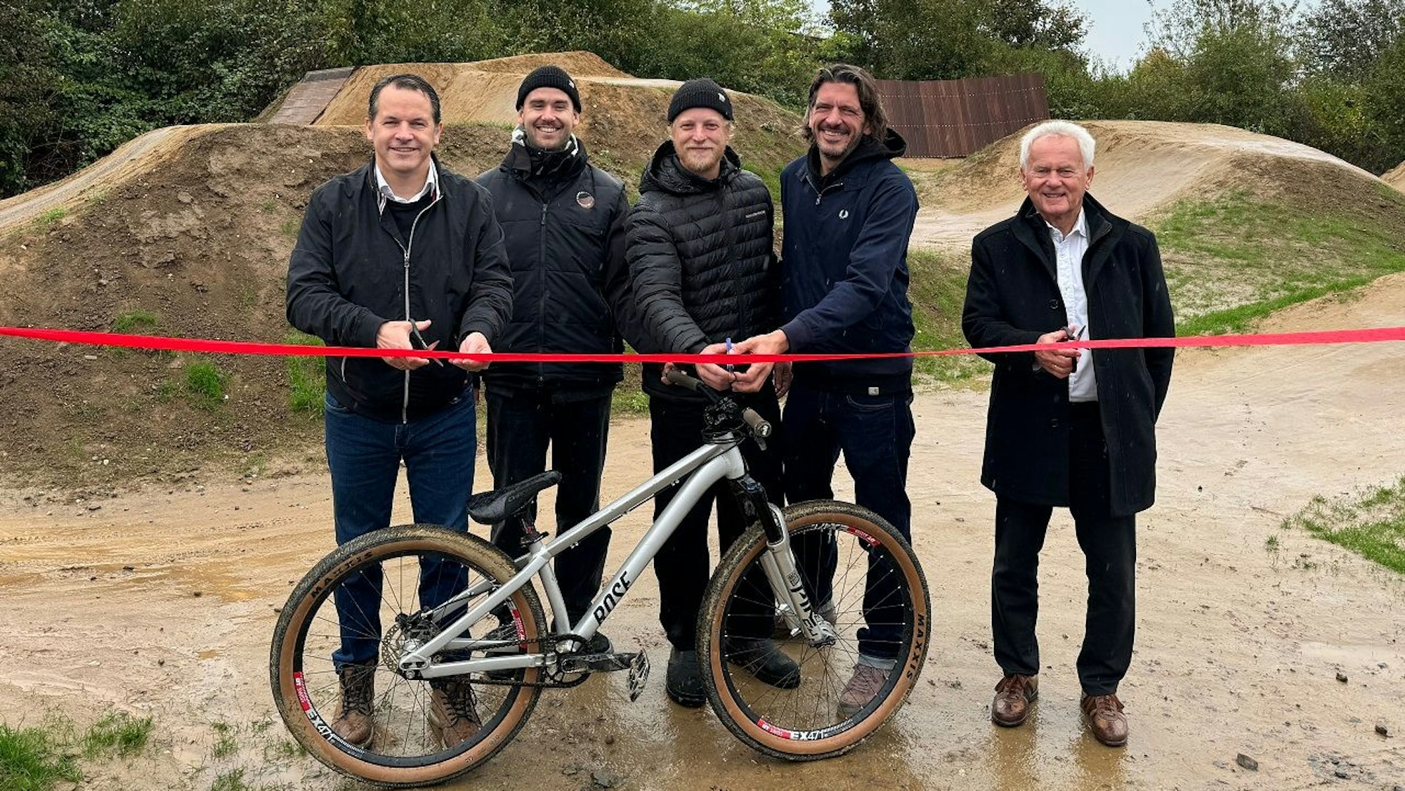 Fünf Männer lächeln in die Kamera, drei sind dabei ein rotes Band zu durchtrennen, vor ihnen steht ein Mountainbike.