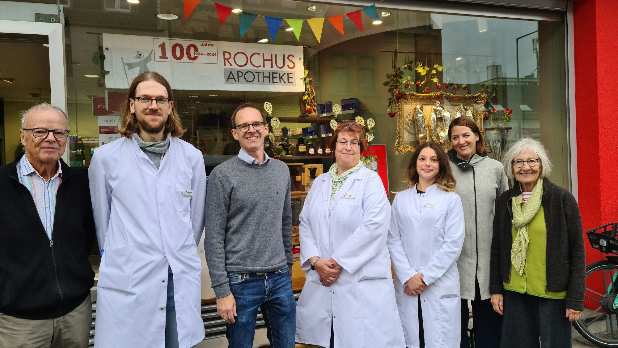 Sieben Menschen, teilweise in weißen Apotheker-Kitteln stehen vor dem Schaufenster der Rochus-Apotheke.