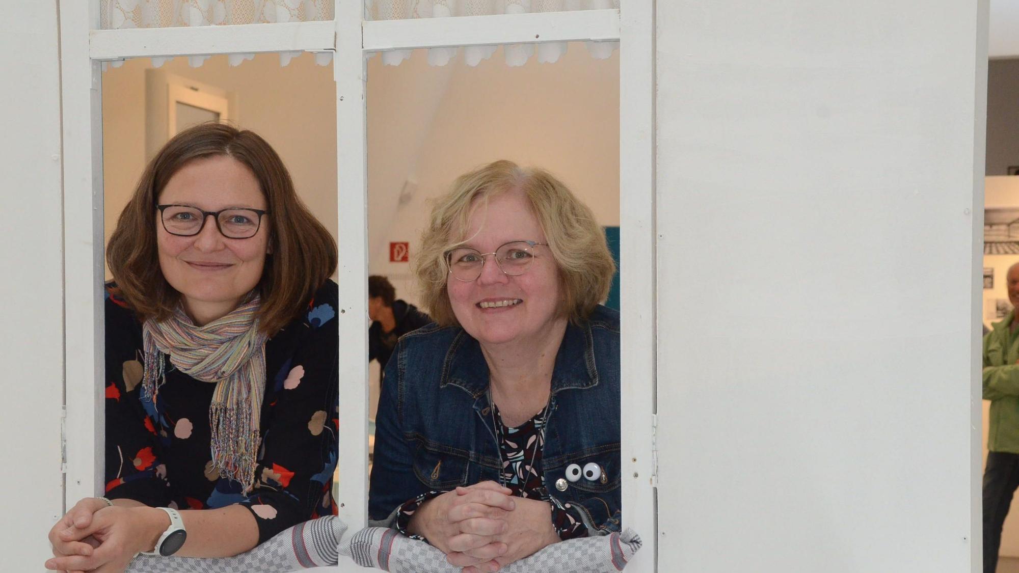 Zwei Frauen schauen durch ein nachgebautes Fenster. Sie haben die Arme auf der gepolsterten Fensterbank abgestützt.