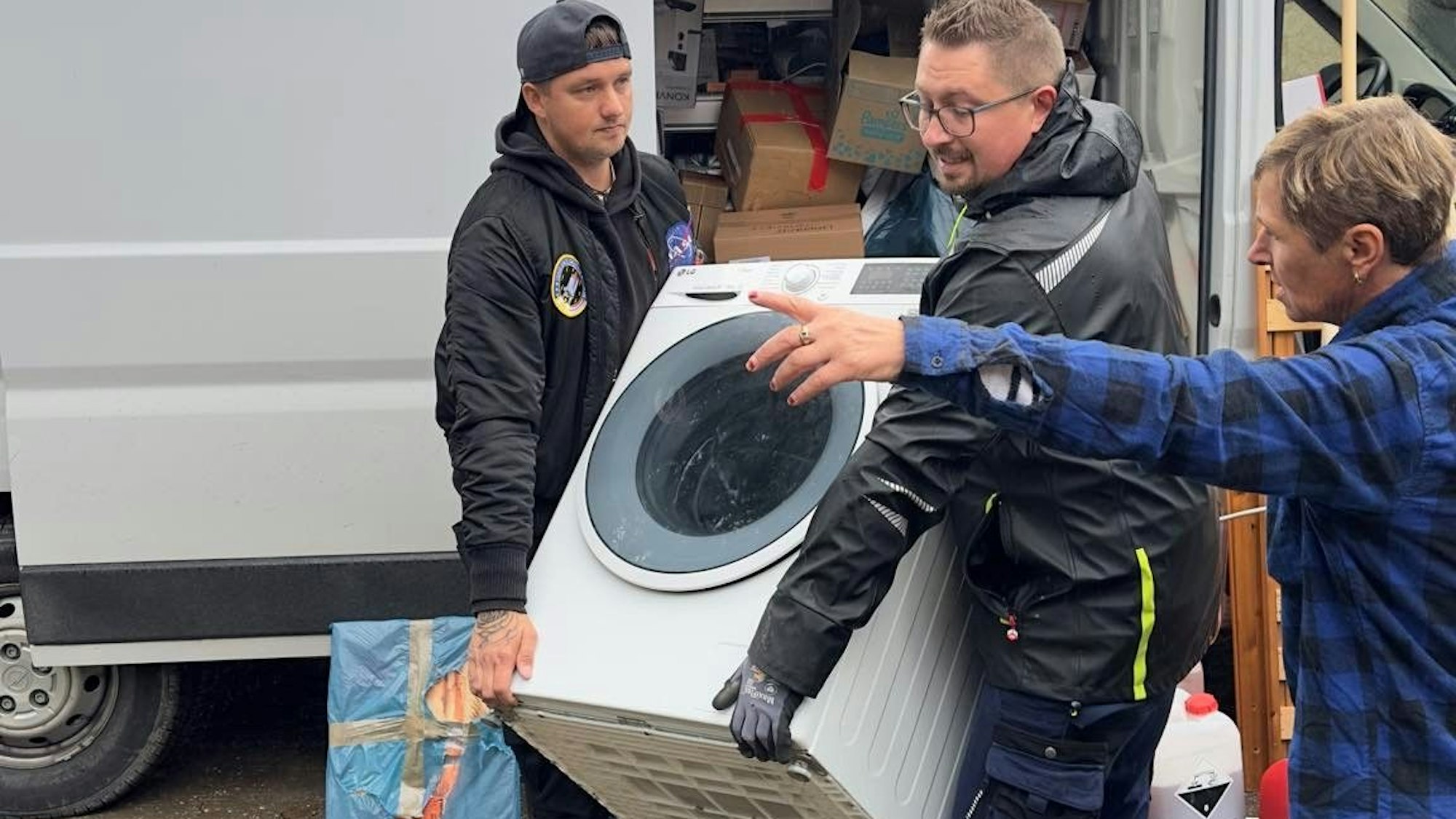 Das Bild zeigt Emil Chrostowska, der mit einem Freund eine Waschmaschine trägt. Eine Frau weist dem Duo den Weg.