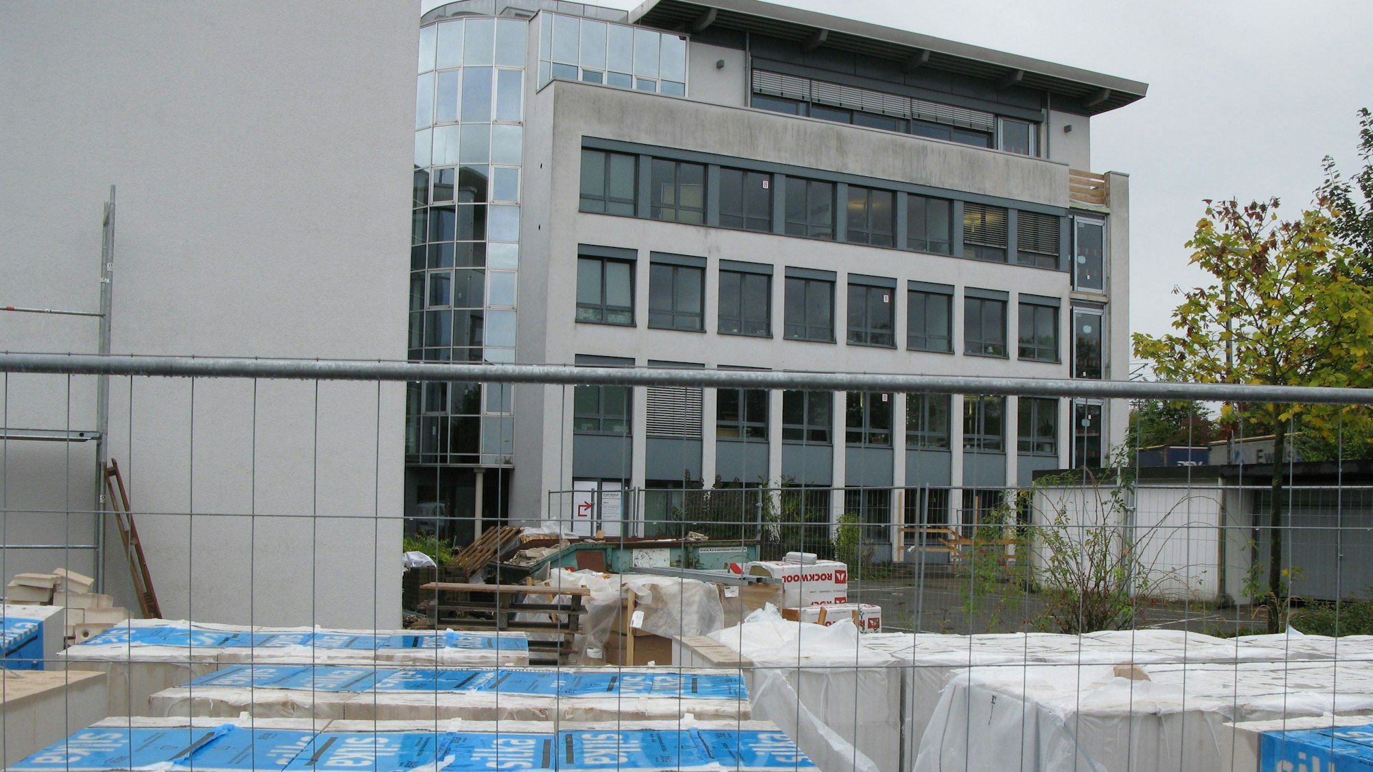 Blick durch einen Bauzaun auf ein fünfstöckiges Bürogebäude, das gerade zu einer Schule umgebaut wird.