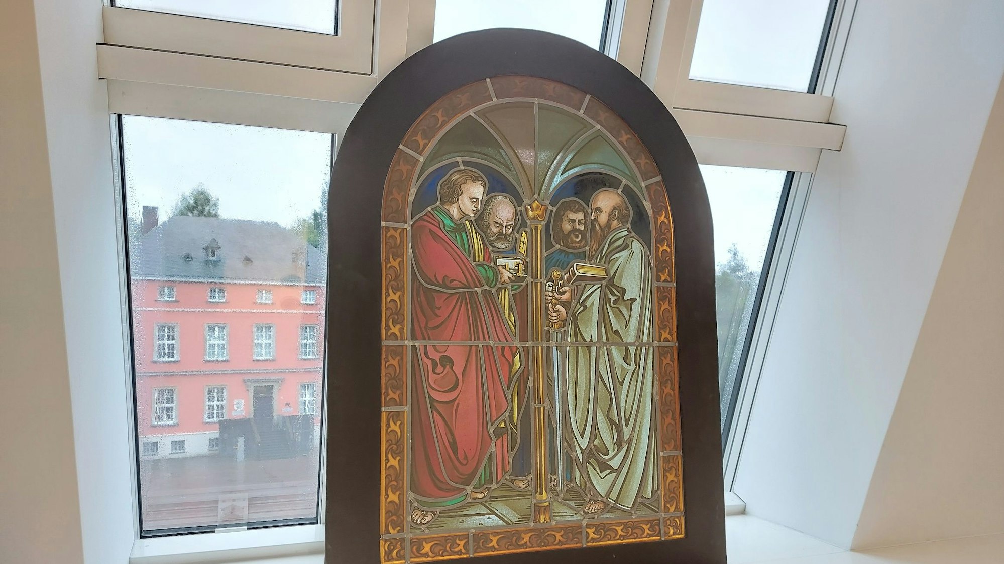 Ein historisches Buntglasfenster vor einem modernen Dachfenster; im Hintergrund ein historisches Herrenhaus mit Sprossenfenstern.