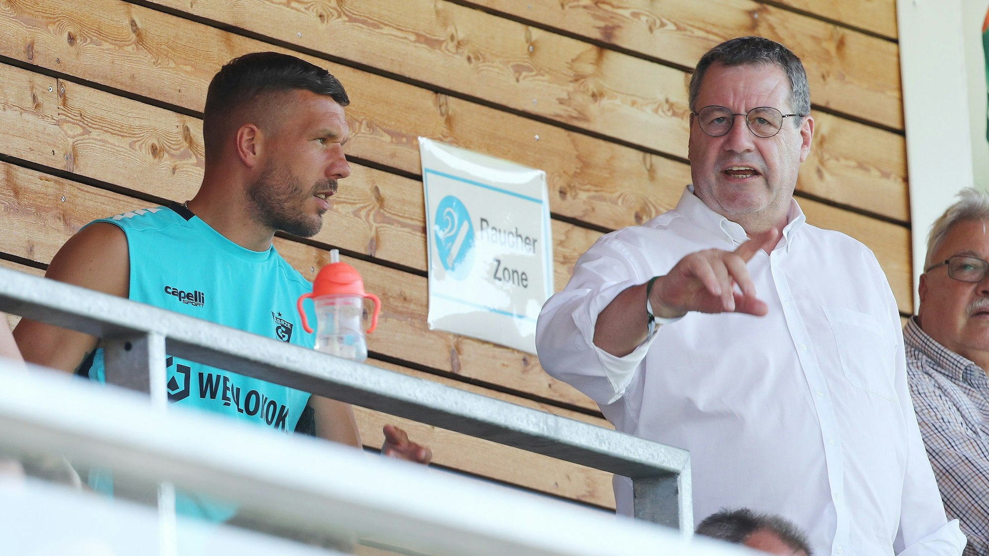 Werner Wolf spricht mit Lukas Podolski auf einer Tribüne.