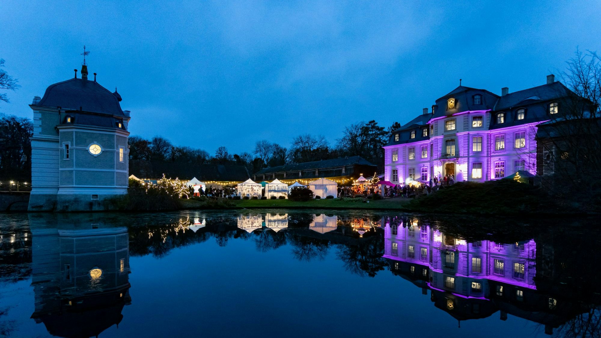 Das Bild zeigt ein beleuchtetes Wasserschloss am Abend, Weihnachtsbuden säumen das Ufer.