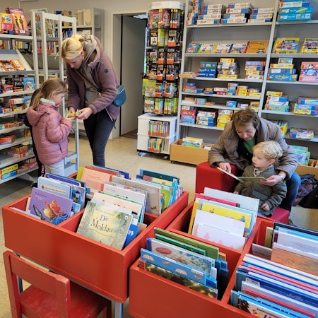 Eine Frau und ihre Tochter stöbern in den Regalen der Bücherei, eine Frau liest einem kleinen Jungen aus einem Buch vor.
