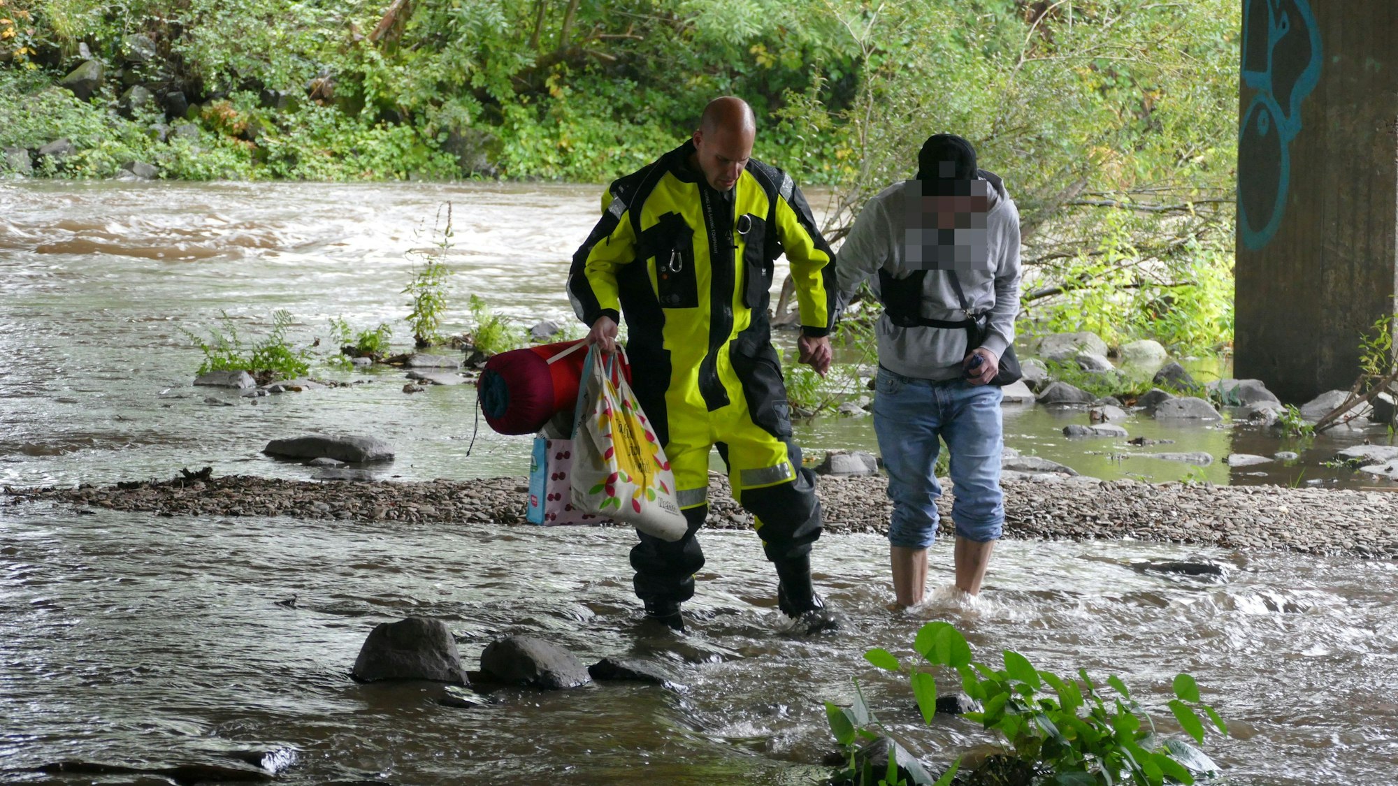 Ein Feuerwehrmann im Überlebensanzug führt einen Mann durch einen Fluss, in der linken Hand hält er zwei Tüten und einen Schlafsack.