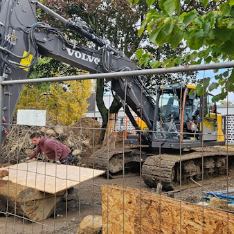 Ein Bagger steht hinter einem Bauzaun vor einer Schule.
