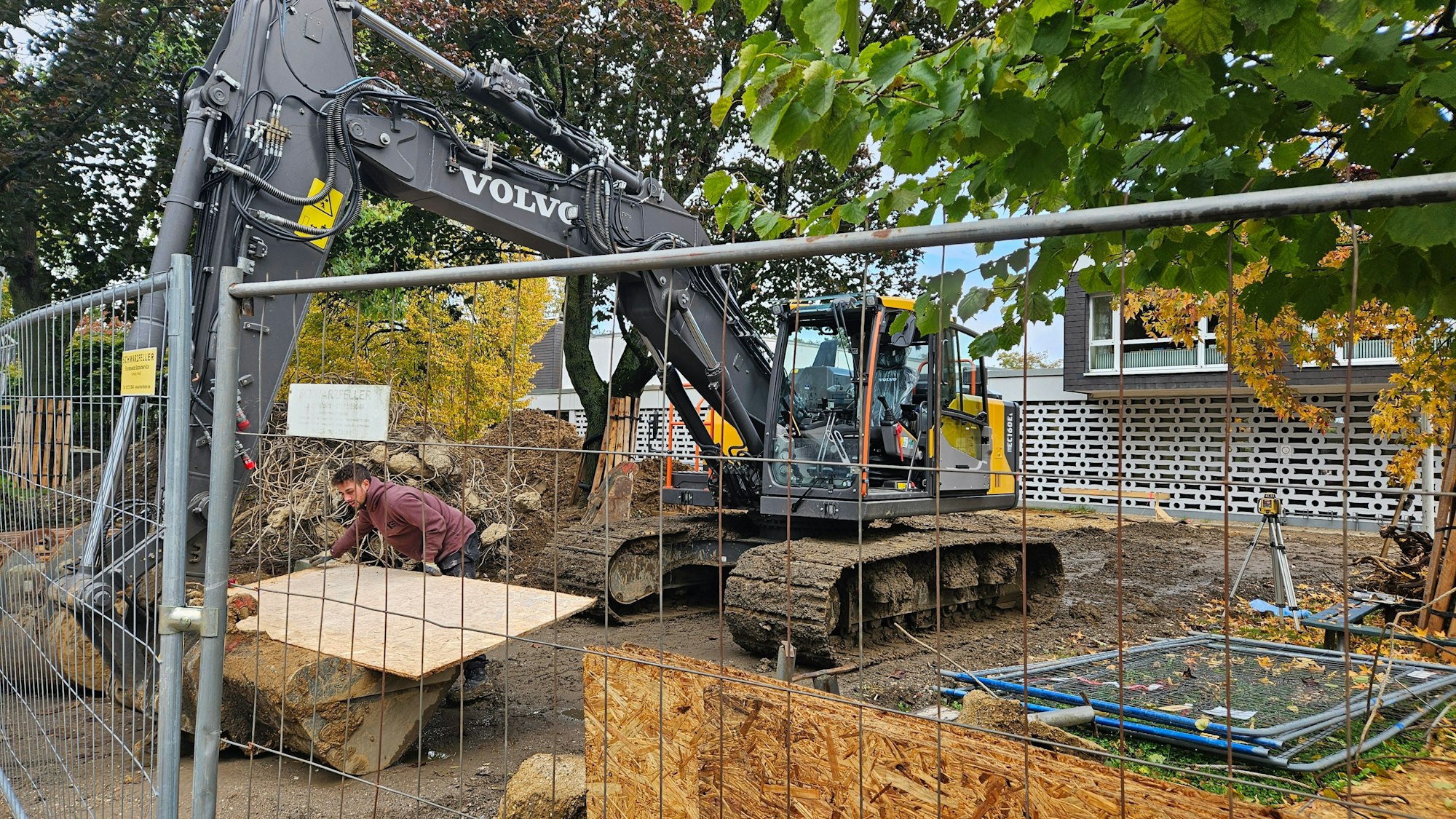 Ein Bagger steht hinter einem Bauzaun vor einer Schule.