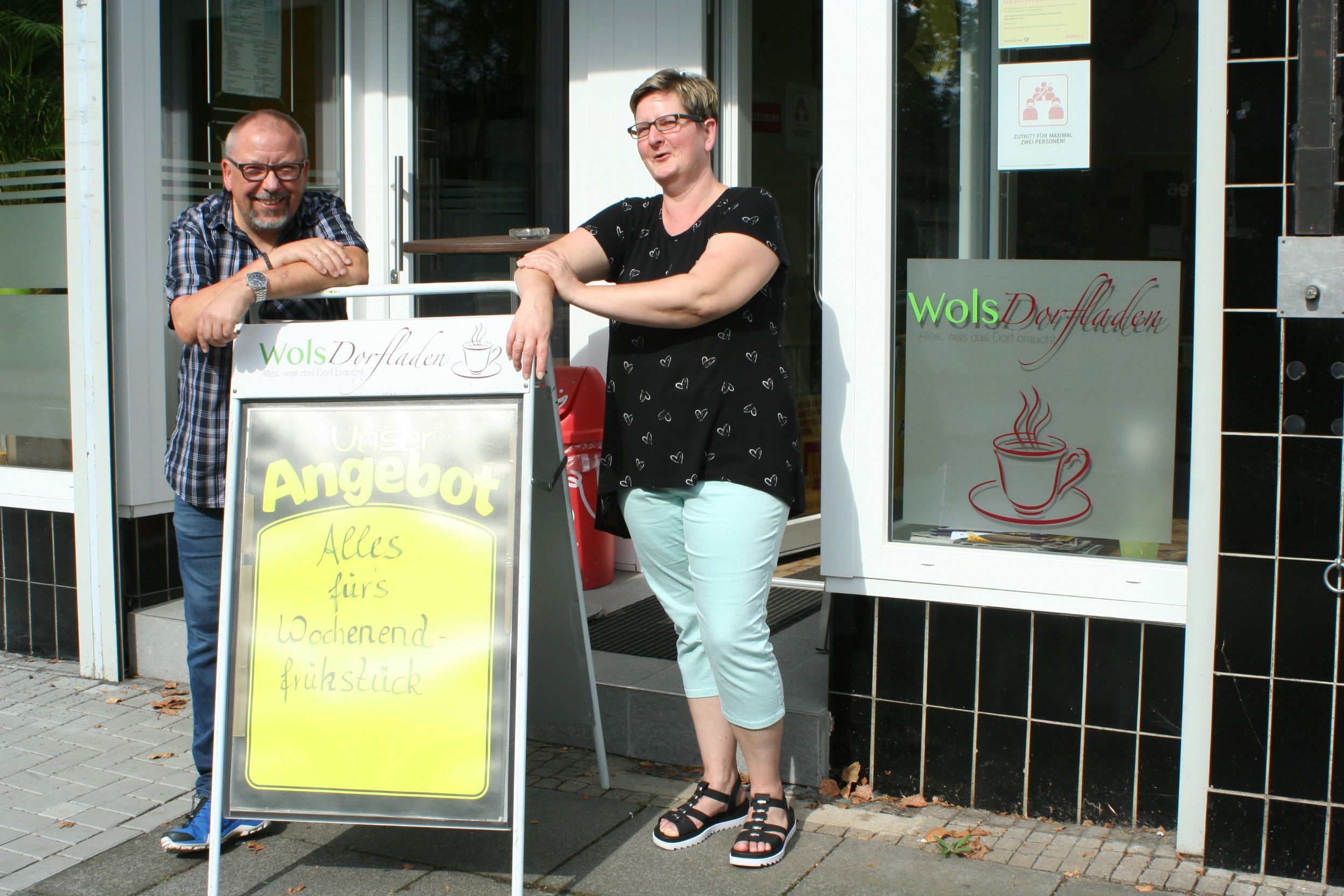 Ein Mann und eine Frau stehen vor einem Ladenlokal mit dem Logo „WolsDorfladen“.