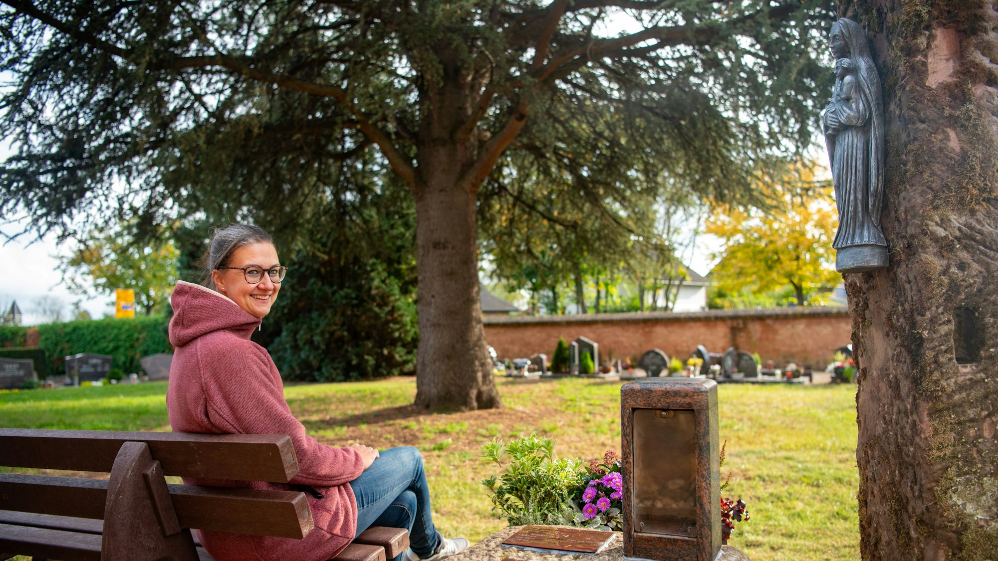 Krystina Antons sitzt auf einer Bank, rechts daneben steht ein Stein mit einem Blumengesteck und einer Halterung für ein Grablicht, rechts daneben steht eine Stele, an der eine Marienfigur hängt.