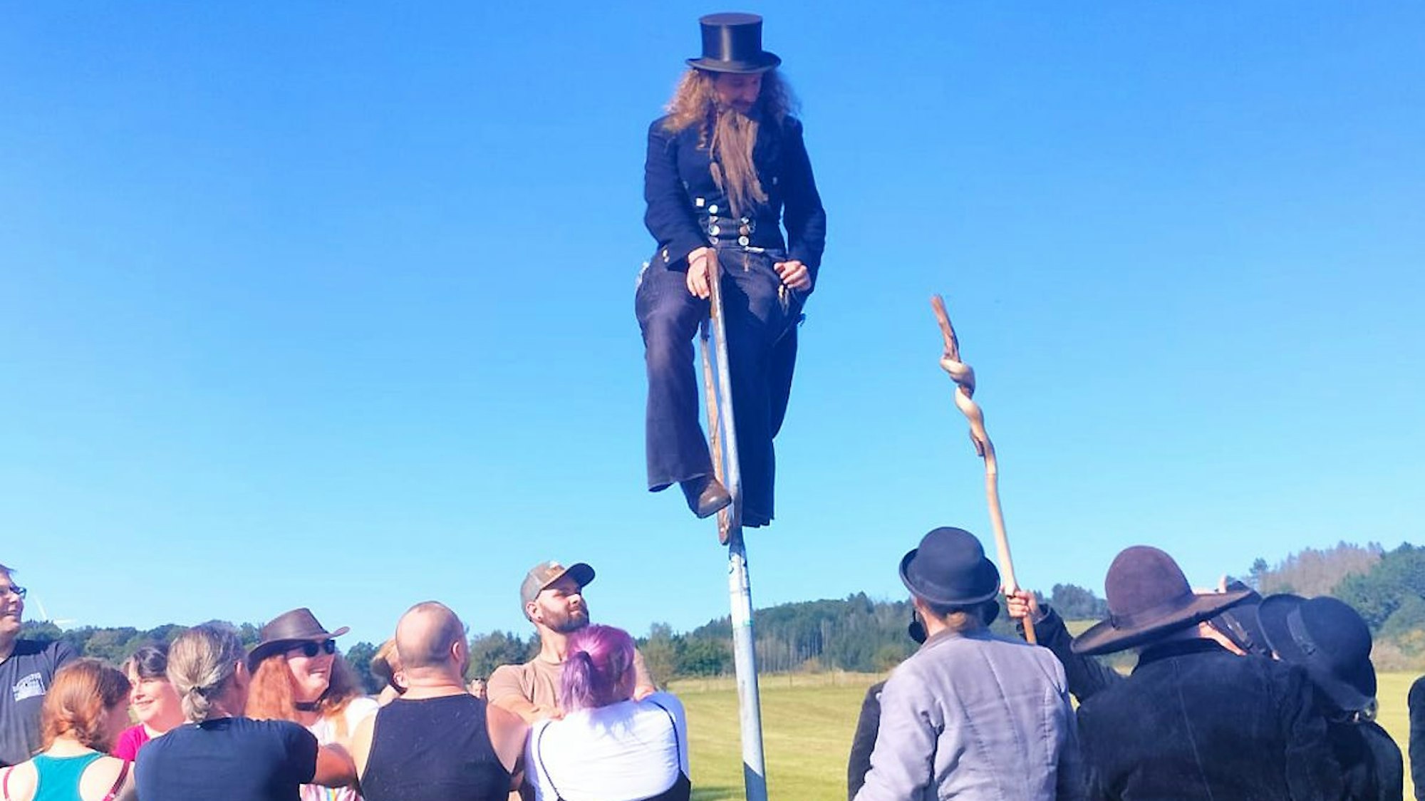 Ein Mann mit Zylinder und angeklebten Bart klettert über ein Ortsschild während ihn Leute anschauen.