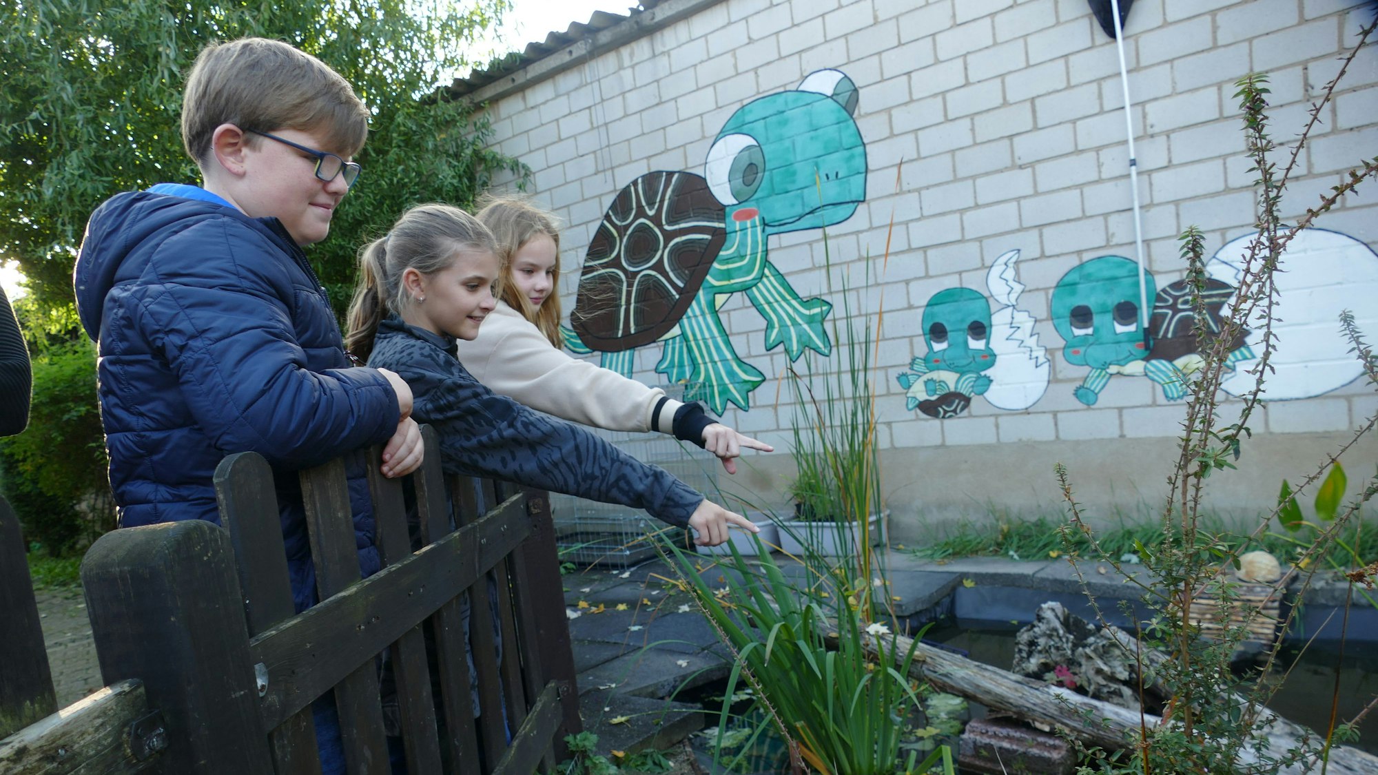 Drei Kinder an einem Holzzaun vor einem Teich.