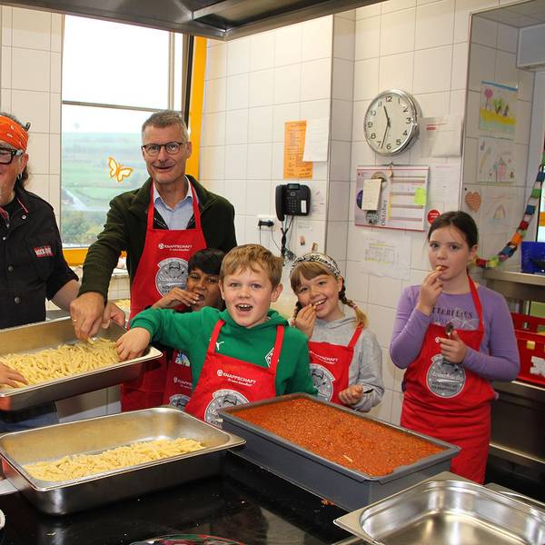 Zwei Männer und eine Gruppe Kinder stehen in einer Küche, in der große Behälter mit Nudeln und einer roten Sauce stehen.
