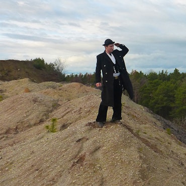 Ein Mann in traditioneller Bekleidung schaut auf einem Hügel in die Ferne. Im Hintergrund befindet sich ein Wald.