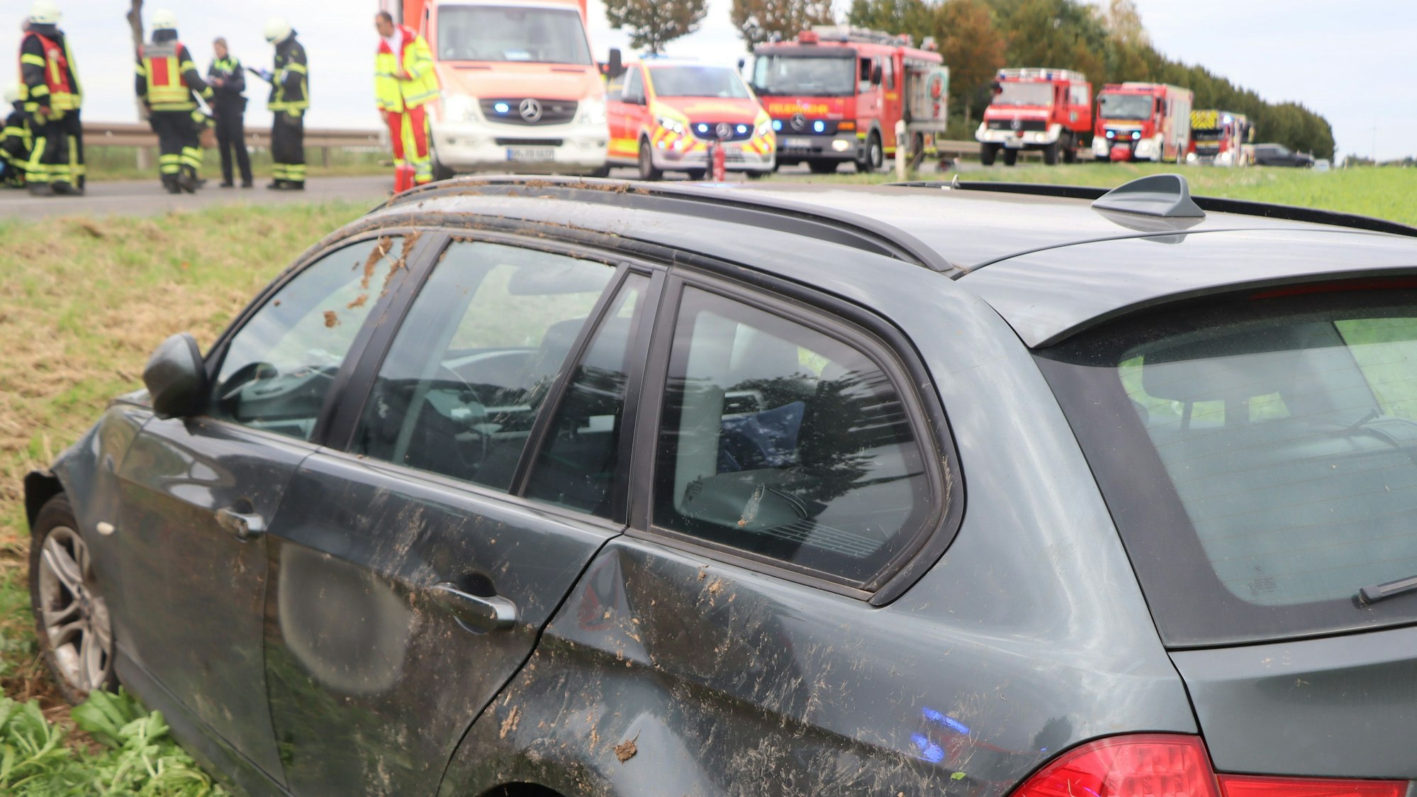 Ein lädierter Wagen mit Beulen und verdreckter Seite steht in einem Feld. Im Hintergrund stehen Feuerwehr und Rettungskräfte mit Fahrzeugen.