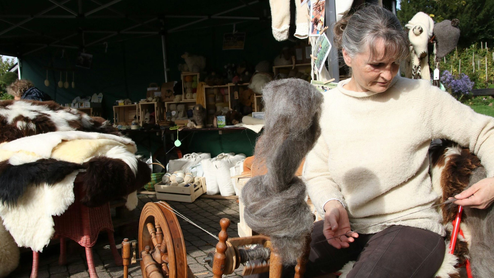 Eine Frau an einem Spinnrad, im Hintergrund ein Stand mit Fellen und Wolle.