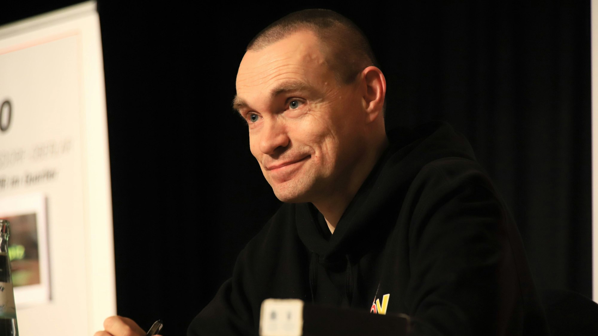 Ein Mann mit millimeterkurz geschnittenen Haaren und schwarzem Pullover.