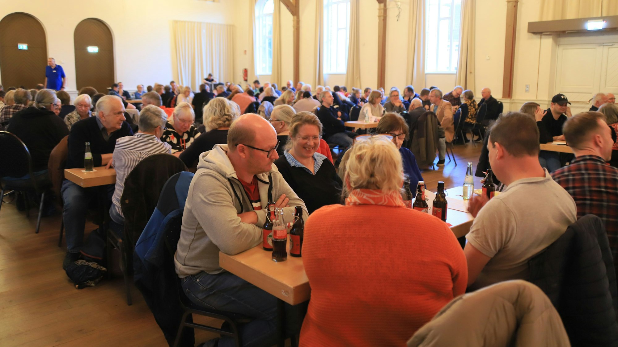 Männer und Frauen sitzen an Tischen in einem vollbesetzten Veranstaltungssaal..