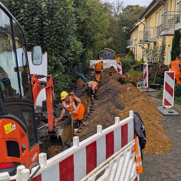Das Foto zeigt einen Bagger vor einer Baugrube, in der Bauarbeiter gerade Glasfaserkabel verlegen.