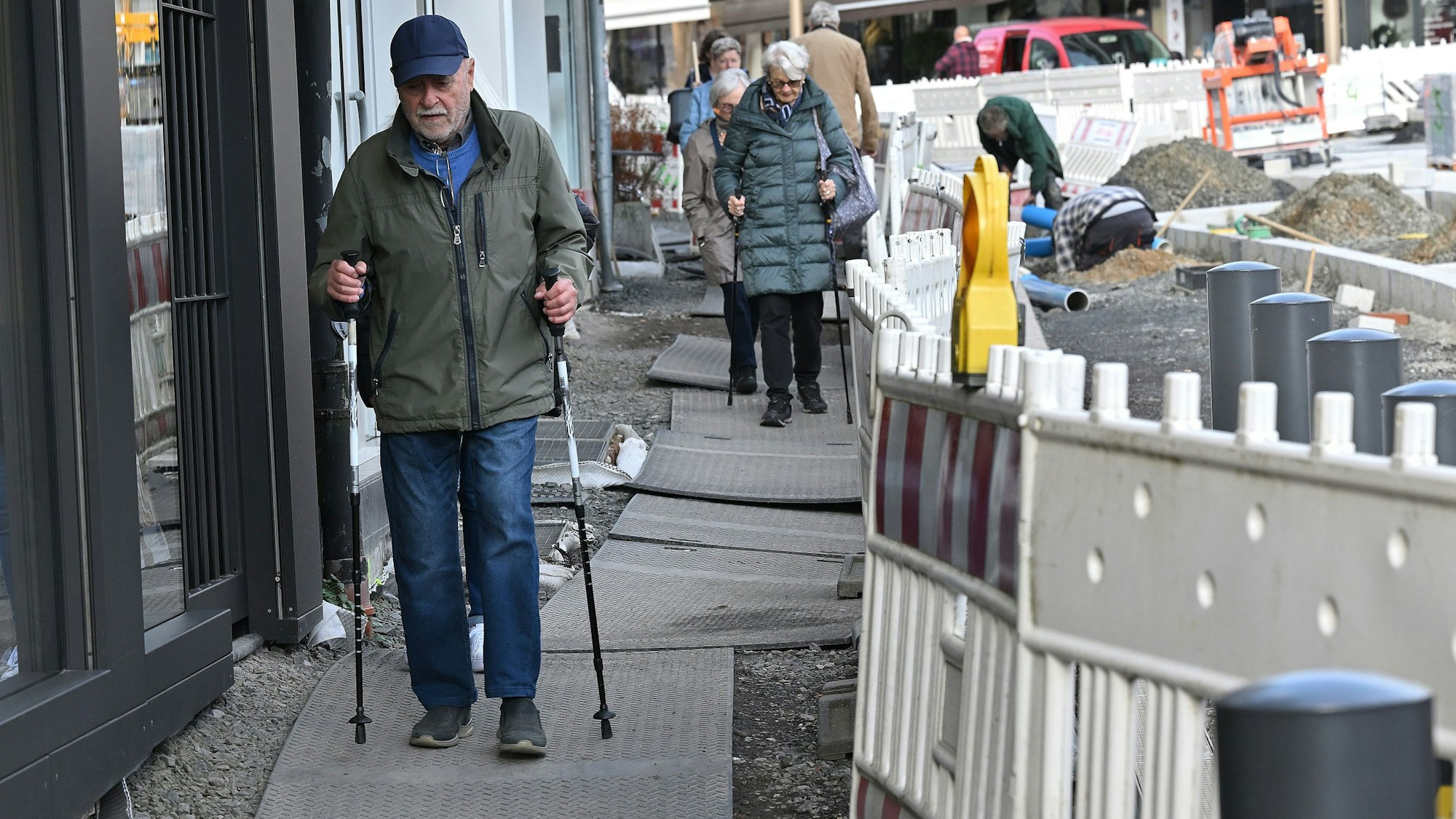 Im Vordergrund geht ein älterer Mann mit zwei Stöcken über Gummimatten. Links sind Geschäftsfassaden, rechts die rot-weißen Absperrungen.